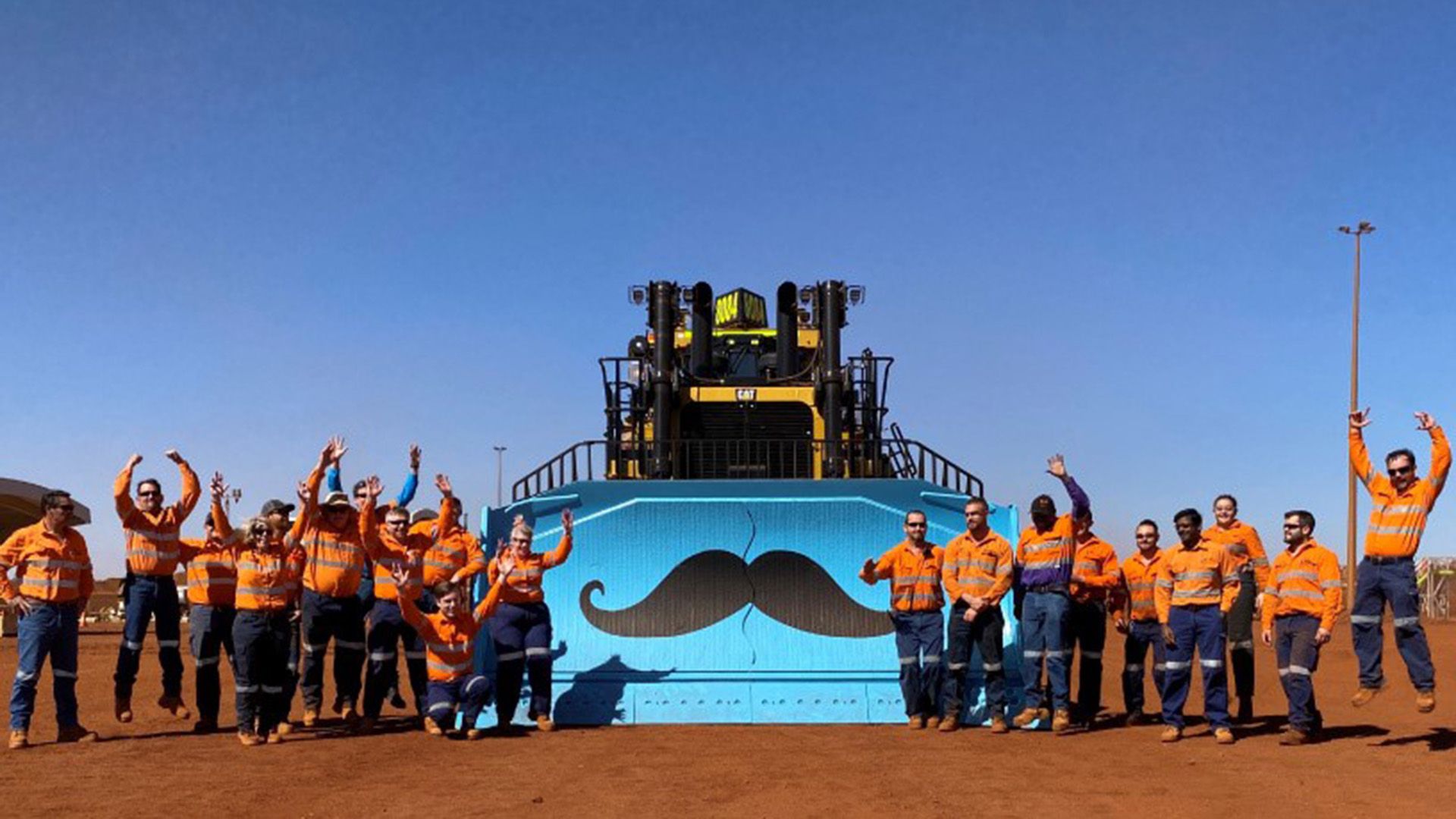 Mining workers in high vis gear, jubilantly standing and jumping before a heavy bulldozer with an emblazoned Movember moustache logo painted on the blade.